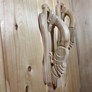 May include: Two light-colored wooden door handles carved into the shape of birds. The handles are mounted vertically on a light-toned wooden surface with visible grain and knots. Each handle features detailed wing and head carvings in natural wood.