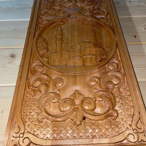 May include: A rectangular wooden carving with a detailed relief of a mosque and ornate floral patterns. The carving is made of light brown wood and features a central oval design with a mosque and minaret.