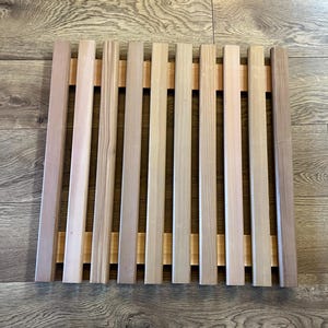 May include: A wooden bath mat with vertical slats of varying shades of light brown and tan. The slats are held together by two horizontal wooden supports, creating a rectangular shape. The wood grain is visible.