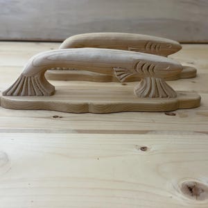 May include: Two light-colored wooden door handles carved into the shape of fish. Each handle features a horizontal body with detailed fin carvings at the base and head, mounted on a flat, elongated wooden plate with rounded edges and screw holes.