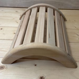 Può includere: Un estensore per la schiena in legno chiaro caratterizzato da una struttura curva e arcuata composta da diverse doghe parallele. La venatura naturale del legno è visibile in tutta la struttura, che presenta una finitura liscia e arrotondata sui bordi superiore e inferiore.