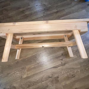May include: A light-colored wooden bench with a rectangular top and angled legs. The bench is constructed with visible wood grain and is approximately 36 inches long. The design is simple and functional, suitable for indoor or outdoor use.