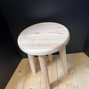 May include: A small, light-colored wooden stool with a round seat and three angled legs. The wood grain is visible on the seat and legs. The stool is designed for seating or as a decorative accent piece.