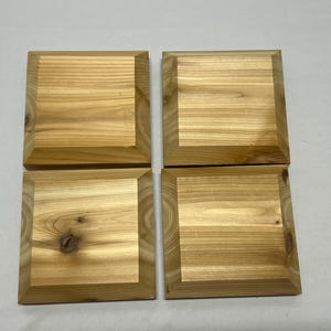 Può includere: Un set di quattro sottobicchieri quadrati in legno disposti in una griglia due per due. Ogni sottobicchiere presenta un motivo a venature del legno naturale marrone chiaro con nodi visibili e un design a bordo smussato, che mostra la texture.