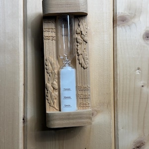 May include: A wooden hourglass with a clear glass bulb containing white sand. The frame features carved oak leaf and acorn motifs. Black markings on the glass and wood indicate 5min, 10min, and 15min intervals. The device is mounted vertically.