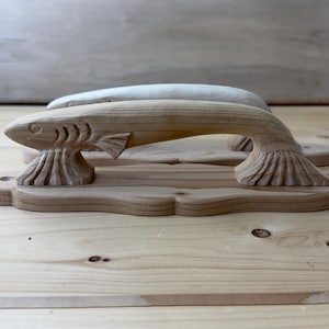 May include: Two light-colored wooden door handles, each carved into the shape of a fish. The handles feature detailed fins and gills, mounted on a flat, scalloped-edge wooden base plate with visible screw holes at each end for mounting purposes.