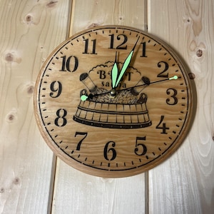 May include: Wooden wall clock with a sauna theme. The clock face is light brown with black numerals and hands. The design includes a sauna bucket and the text "BÄT sauna". The clock is mounted on a light wood wall.