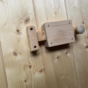 Könnte beinhalten: Ein hölzerner Riegel mit Knauf und rechteckigem Verschluss. Der Riegel ist aus hellfarbenem Holz gefertigt und an einer Holzoberfläche befestigt. Das Design ist einfach und rustikal, geeignet für eine Tür oder ein Tor.