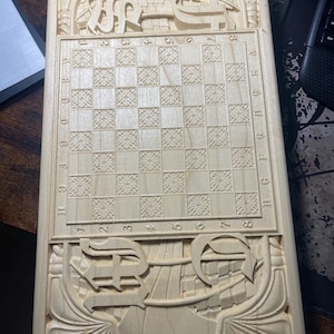 Puede incluir: Un tablero de ajedrez de madera de color claro con intrincadas tallas. El tablero presenta un patrón de tablero de ajedrez, bordes decorativos y tallas ornamentadas en la parte superior e inferior. El tablero es rectangular y parece estar hecho de madera de color claro.