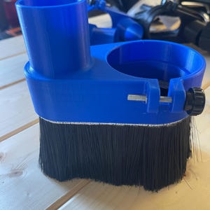 May include: A blue CNC router dust shoe with black bristles. The dust shoe has a cylindrical opening and a clamp with a screw for attachment. The bristles are designed to contain dust and debris during routing operations.