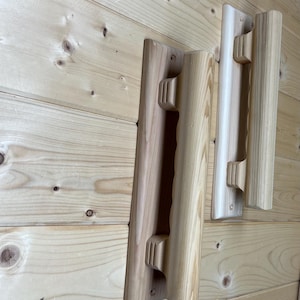 May include: Three wooden door handles with a natural finish are mounted on a light-colored wooden wall. The handles have a rectangular base and a cylindrical grip with a carved design. The wood grain is visible.