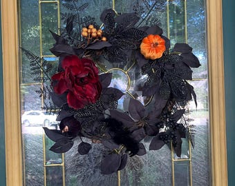 May include: A black and burgundy Halloween wreath with black leaves, a red rose, and an orange pumpkin. The wreath is hanging on a glass door with a gold frame.