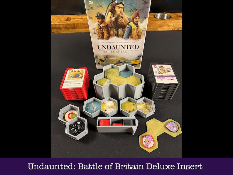 May include: A board game insert for "Undaunted: Battle of Britain". The image displays the game box, stacks of cards, hexagonal game tiles, and other components in red, grey, and yellow. The box art features illustrations of pilots and aircraft.