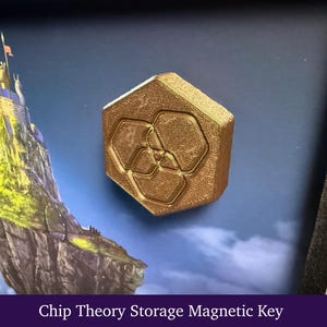 Könnte beinhalten: Ein goldfarbener, sechseckiger magnetischer Schlüssel mit strukturierter Oberfläche und einem zentralen geometrischen Emblem. Der Schlüssel ist auf einem dunkelblauen Hintergrund mit einer Burg auf einem Felsen und dem Text Chip Theory Storage Magnetic Key montiert.