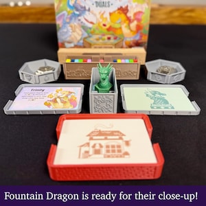 May include: A board game setup featuring a green dragon figurine, cards, and game pieces. The game box is visible in the background. The text "Fountain Dragon is ready for their close-up!" is at the bottom.