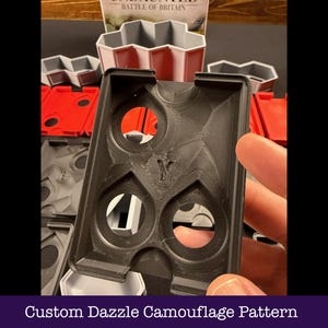 May include: Black plastic game organiser with a custom dazzle camouflage pattern. The organiser has multiple circular cutouts and a stylized "Y" emblem. Other game components in red, grey, and white are visible in the background.