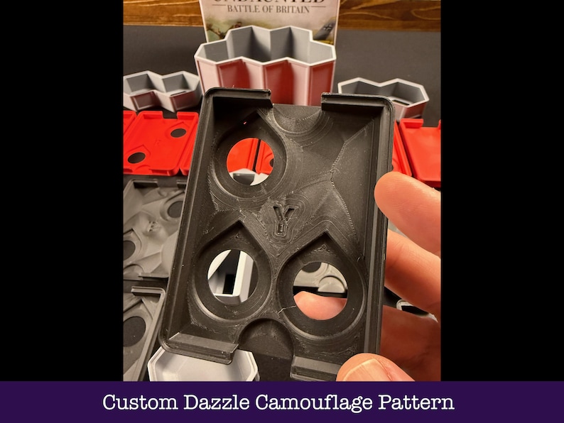 May include: Black plastic game organiser with a custom dazzle camouflage pattern. The organiser has multiple circular cutouts and a stylized "Y" emblem. Other game components in red, grey, and white are visible in the background.