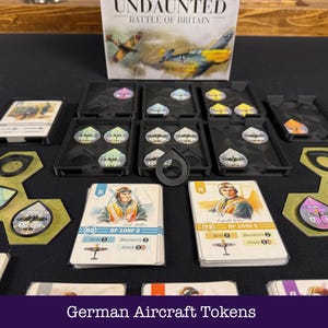 May include: A board game setup with the "Undaunted: Battle of Britain" box visible. The image shows game components such as cards with pilot illustrations, aircraft tokens, and black trays. Text at the bottom reads "German Aircraft Tokens".