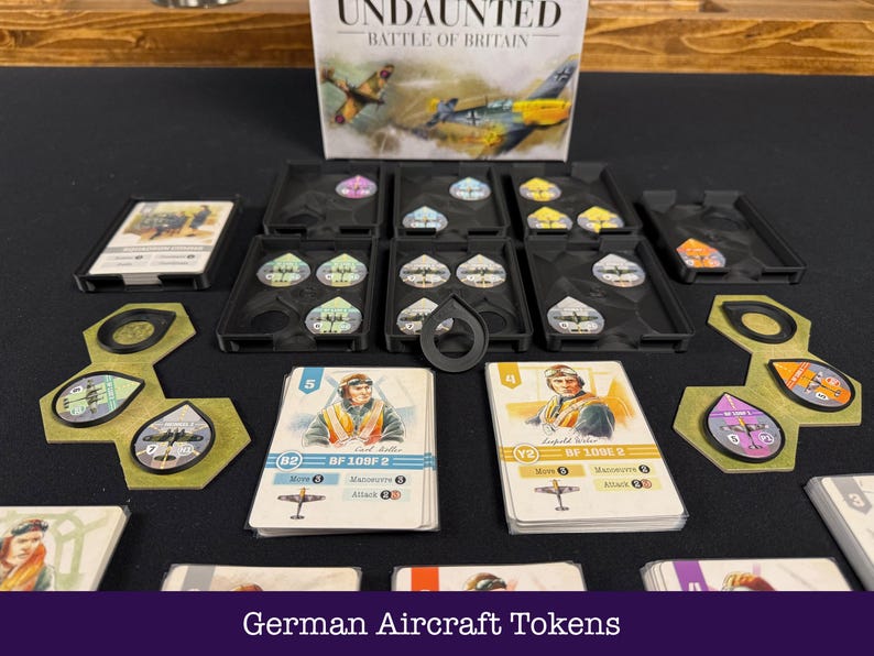 May include: A board game setup with the "Undaunted: Battle of Britain" box visible. The image shows game components such as cards with pilot illustrations, aircraft tokens, and black trays. Text at the bottom reads "German Aircraft Tokens".