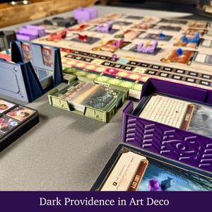 Puede incluir: Un juego de mesa con un tablero grande, varias cartas, fichas y piezas de jugador de plástico de colores. Varios tarjeteros impresos en 3D en azul, verde y morado están dispuestos sobre la mesa, conteniendo mazos de cartas con texto.