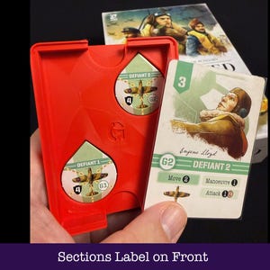 May include: A red plastic game component holder with two circular cutouts, each containing a game piece with a plane graphic. A game card with a pilot illustration and the text "DEFIANT 2" is held next to the holder. The text "Sections Label on Front" is at the bottom.