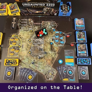 Peut inclure: Mise en place d'un jeu de soci&eacute;t&eacute; avec un plateau de jeu d&eacute;taill&eacute;, des cartes et divers &eacute;l&eacute;ments de jeu. Le jeu comprend des porte-cartes jaunes et bleus, des d&eacute;s et une bo&icirc;te avec le titre du jeu "Undaunted 2200 Callisto". Le texte "Organized on the Table!" est en bas.