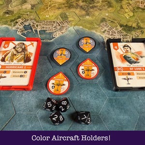 May include: A board game setup with aircraft cards in red and black holders, and circular and teardrop-shaped tokens. Black dice with white numbers are scattered around the game pieces. The text "Color Aircraft Holders!" is at the bottom.