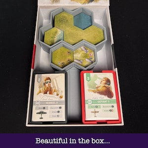May include: A board game box with a grey interior, containing game pieces and cards. The box features a hexagonal terrain piece, two card holders with game cards, and the text "Beautiful in the box..." at the bottom.
