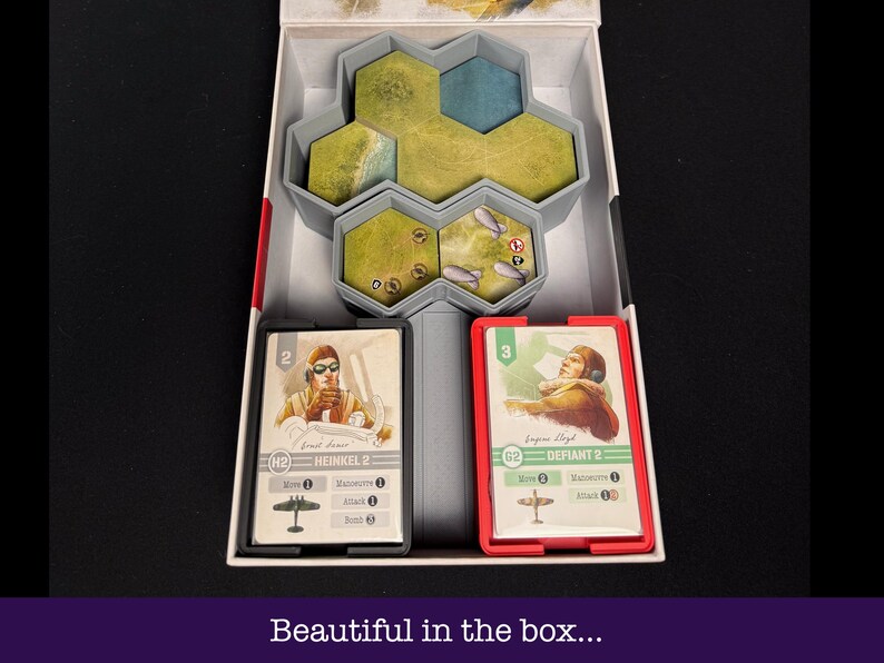May include: A board game box with a grey interior, containing game pieces and cards. The box features a hexagonal terrain piece, two card holders with game cards, and the text "Beautiful in the box..." at the bottom.