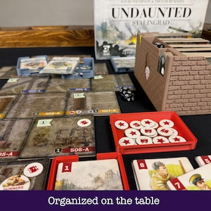 Può includere: Un set da gioco da tavolo Undaunted Stalingrad con un organizzatore di carte marrone effetto mattoni, un vassoio rosso pieno di gettoni circolari bianchi con stelle rosse, varie tessere mappa quadrate e carte personaggio illustrate.
