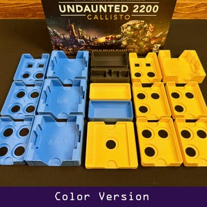 Peut inclure: Une bo&icirc;te de jeu de soci&eacute;t&eacute; intitul&eacute;e "Undaunted 2200 Callisto" avec divers organisateurs de composants de jeu en plastique bleu, jaune et noir. Les organisateurs sont dispos&eacute;s sur une surface sombre. Le texte "Color Version" est en bas.
