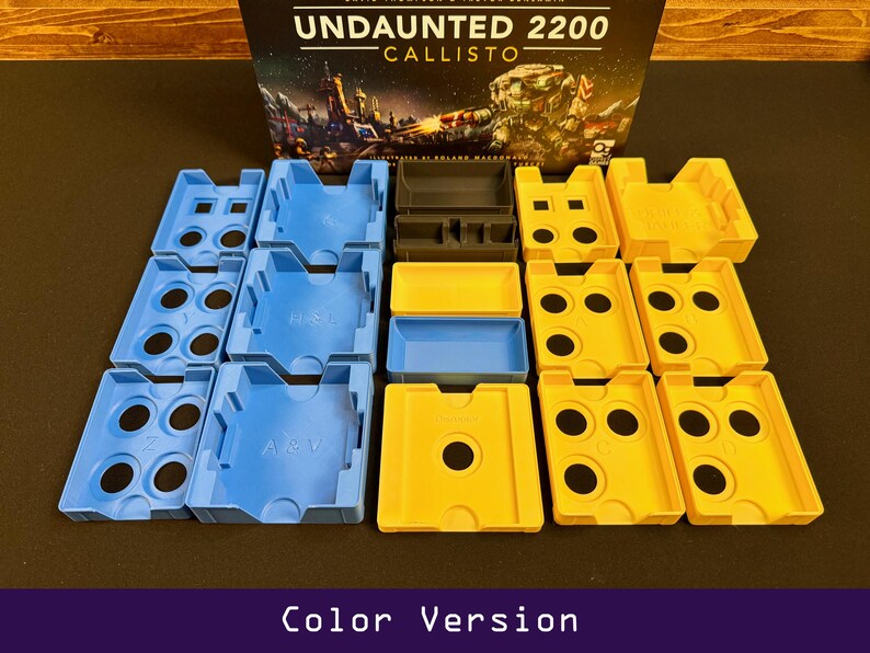 Peut inclure: Une bo&icirc;te de jeu de soci&eacute;t&eacute; intitul&eacute;e "Undaunted 2200 Callisto" avec divers organisateurs de composants de jeu en plastique bleu, jaune et noir. Les organisateurs sont dispos&eacute;s sur une surface sombre. Le texte "Color Version" est en bas.
