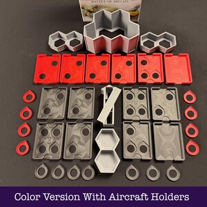 May include: A selection of grey, red, and white game components, including aircraft holders, displayed on a dark surface. The set features diverse shapes and sizes, such as rectangular, hexagonal, and circular pieces. The text "Color Version With Aircraft Holders" is at the bottom.