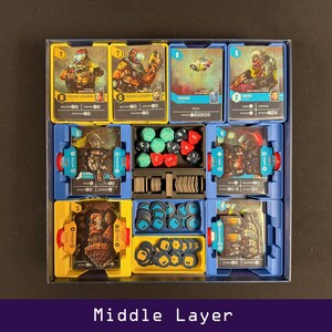 Peut inclure: Un organisateur de jeu avec plusieurs compartiments remplis de cartes de jeu, de d&eacute;s et de jetons. Les cartes pr&eacute;sentent des illustrations de personnages et de v&eacute;hicules. L'organisateur est bleu et jaune, avec le texte "Middle Layer" en bas.