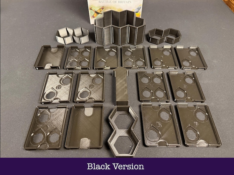 May include: An assortment of black game components, featuring rectangular trays with circular apertures, hexagonal forms, and a concertina-style structure. The text "Black Version" is at the base of the image. The pieces are displayed on a dark surface.