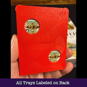 May include: A vibrant red game tray with two circular apertures displaying game pieces. The tray is marked "Section G" and "v17". The text "All Trays Labeled on Back" is at the bottom. The tray is held in a hand.