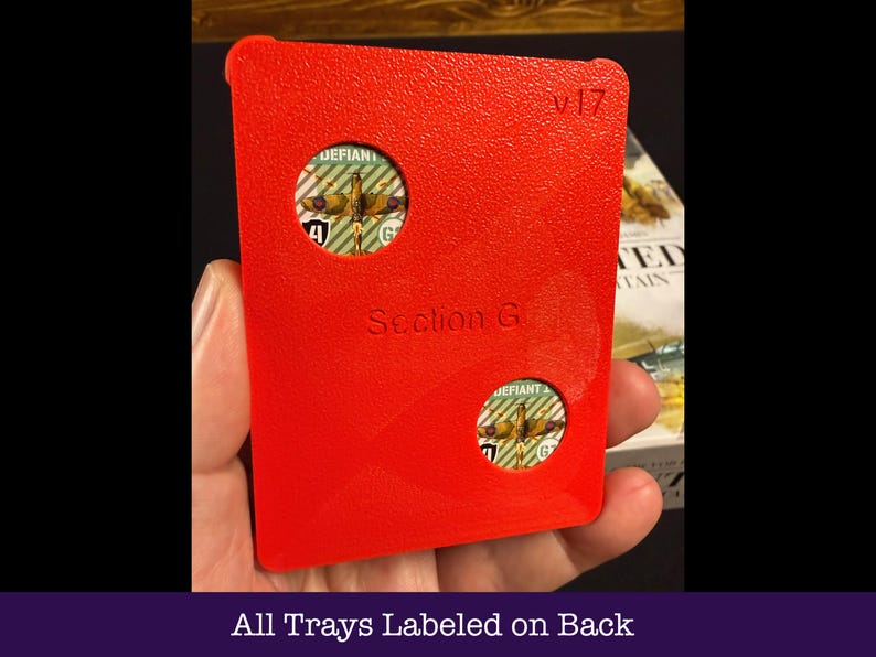 May include: A vibrant red game tray with two circular apertures displaying game pieces. The tray is marked "Section G" and "v17". The text "All Trays Labeled on Back" is at the bottom. The tray is held in a hand.