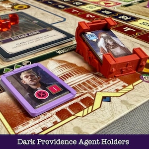 Op de afbeelding: Een bordspelopstelling met kaarten, rode dobbelstenen en agenthouders. Het spelbord toont een kaart met verschillende locaties. De agenthouders zijn rood en paars, ontworpen om kaarten met karakterafbeeldingen te bevatten. De tekst "Dark Providence Agent Holders" staat onderaan.