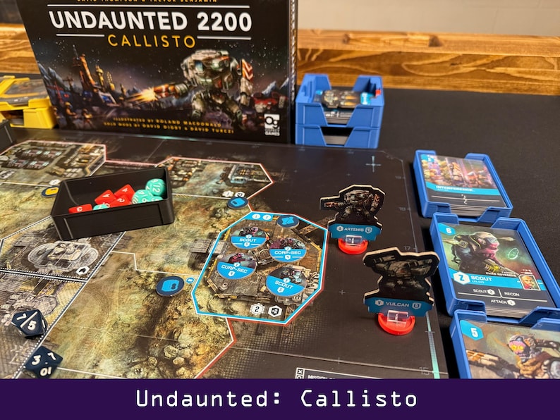 Peut inclure: Mise en place du jeu de soci&eacute;t&eacute; "Undaunted: Callisto". Le plateau de jeu pr&eacute;sente une carte d&eacute;taill&eacute;e avec des pi&egrave;ces, notamment des cartes, des d&eacute;s et des figurines miniatures. La bo&icirc;te de jeu et les conteneurs de rangement bleus sont &eacute;galement visibles.