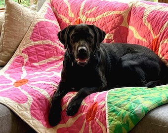 May include: A quilted throw blanket featuring a large-scale floral pattern in vibrant shades of pink, cream, and green. The blanket has a diamond-stitched texture and is draped over a gray sofa, with a black dog resting on top of the fabric surface.