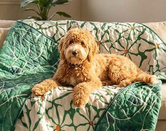 Puede incluir: Una manta acolchada con un patrón repetitivo de flores blancas similares a margaritas con centros naranjas sobre un fondo verde, colocada sobre un sofá beige con un perro doodle de color marrón dorado descansando sobre la tela en una sala.