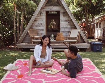 May include: A large rectangular picnic blanket featuring a cream-colored base with a repeating pattern of oversized, quilted pink flowers with orange centers. The blanket is spread out on a grassy lawn in front of a wooden A-frame cabin structure.