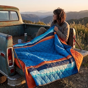 May include: A quilted blanket featuring horizontal stripes in shades of blue, orange, and white. The blanket has a diamond-stitched pattern and is draped over the tailgate of a vintage pickup truck, showcasing a vibrant, multi-colored design.