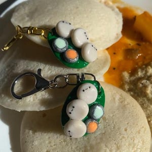 Puede incluir: Dos llaveros con un diseño de vaina de guisantes verde, cada uno con cuentas blancas, naranjas y verdes con pequeñas caras. Los llaveros están colocados sobre idli, un desayuno del sur de la India, con sambar.