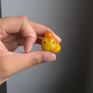 May include: A small, yellow and orange, handmade polymer clay charm shaped like a cartoon animal with a round head, two small eyes, and a small mouth. The charm has a small loop at the top for attaching to a necklace or bracelet.