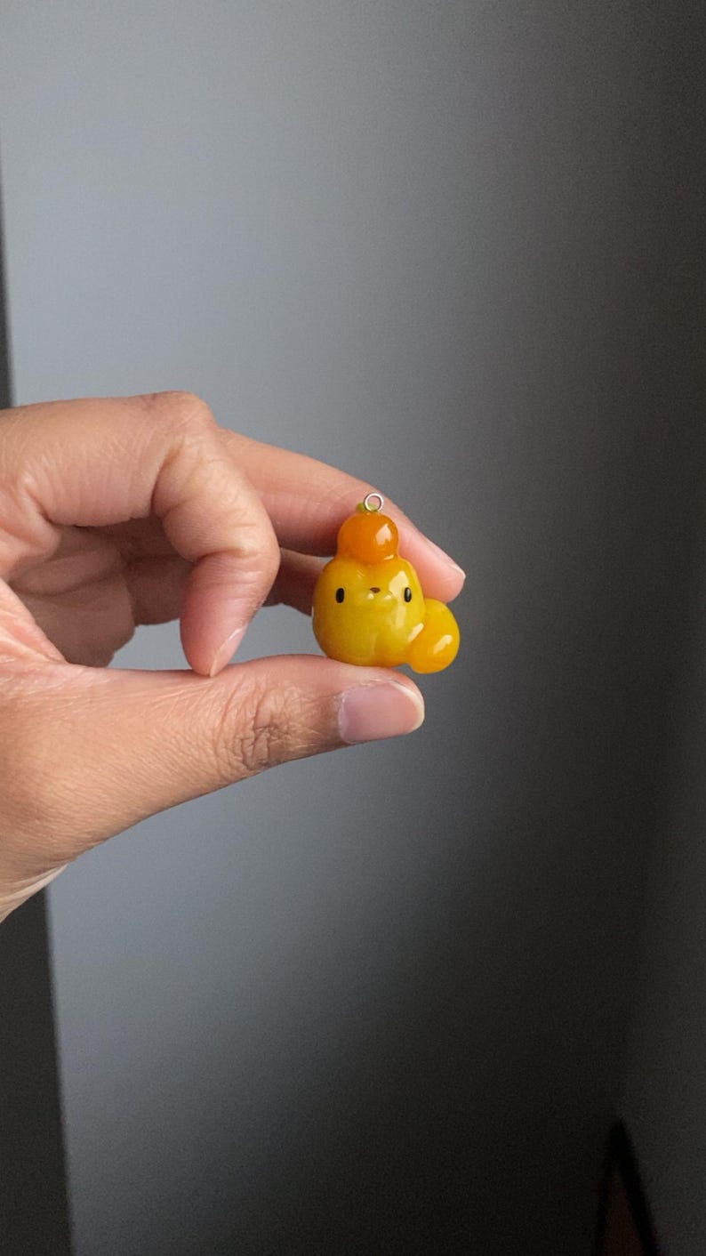 May include: A small, yellow and orange, handmade polymer clay charm shaped like a cartoon animal with a round head, two small eyes, and a small mouth. The charm has a small loop at the top for attaching to a necklace or bracelet.