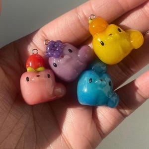 May include: Four colorful, small, handmade, resin charms with cute faces. Each charm has a different color and a different topping. The charms are pink, purple, blue, and yellow.