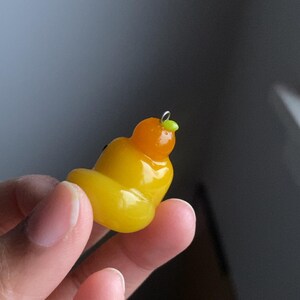 May include: A yellow and orange polymer clay charm shaped like a cartoon character with a green leaf on top. The charm is hanging from a silver loop.