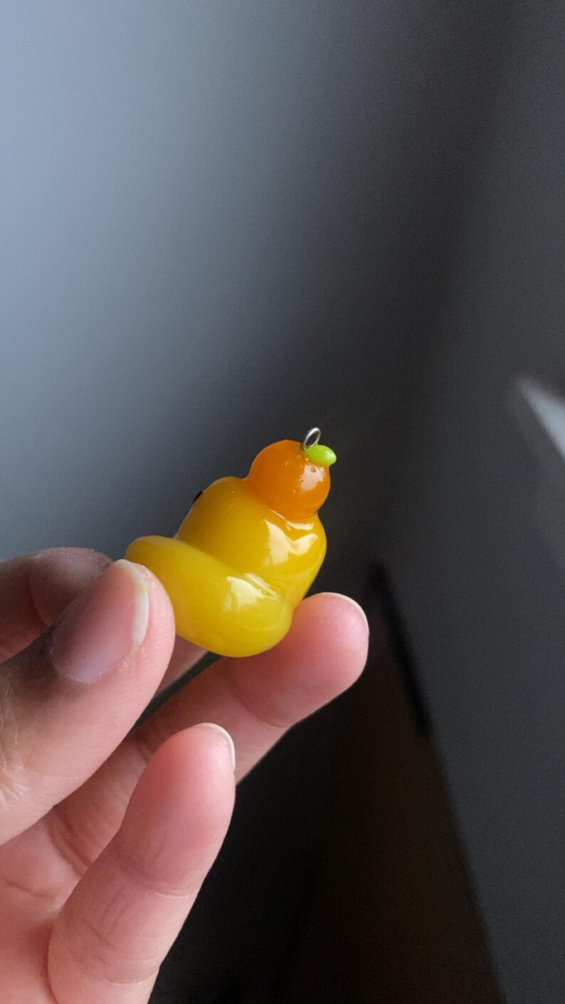 May include: A yellow and orange polymer clay charm shaped like a cartoon character with a green leaf on top. The charm is hanging from a silver loop.