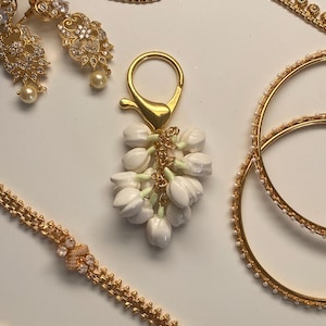 May include: Gold-toned jewelry set featuring a floral charm, earrings, a necklace, and bangles. The charm is a cluster of white flowers with a gold clasp. The earrings and necklace have clear stone accents.